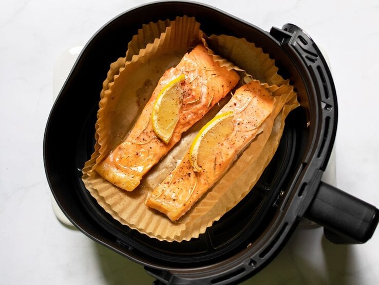 Zwei goldbraun gebackene Lachsfilets mit Zitronenscheiben und Gewürzen liegen auf Backpapier in einem schwarzen Airfryer.