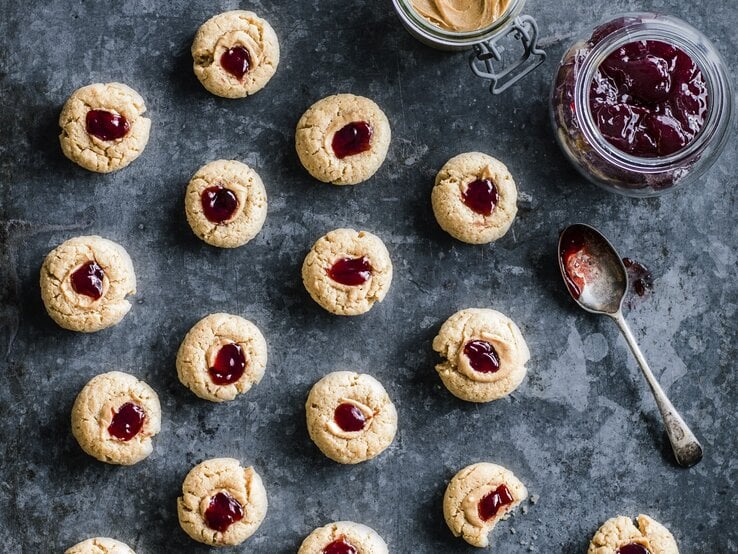 Kekse mit Marmelade und Erdnussbutter auf grauem Hintergrund, rechts ein Glas gefüllt mit Marmelade.