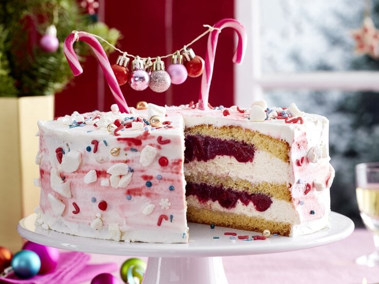 Zuckerstangen-Torte auf einem weißen Tortenteller auf einer rosa Tischdecke dekoriert mit kleinen Weihnachtskugeln. Links hinten auf dem Foto ein kleiner Tannenbaum.