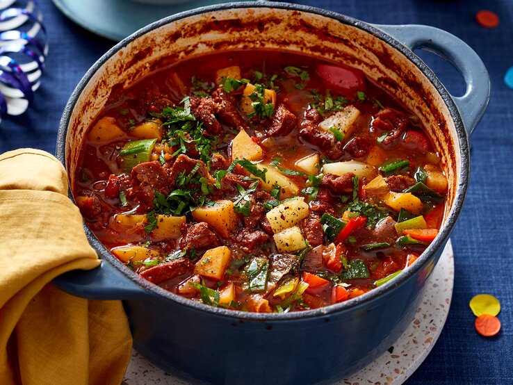 Ofen-Gulaschsuppe in einem blauen Topf auf blauem Hintergrund. Links auf dem Foto ein gelbes Geschirrtuch.