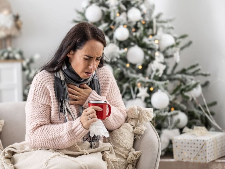 Eine dunkelhaarige Frau in einem rosa Strickpullover sitzt krank auf dem Sofa, hält eine Tasse Tee und presst die Hand an die Brust.