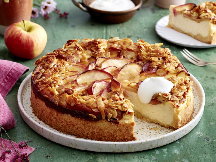 Apfel-Quark-Bienenstich auf einem weißen Teller auf grünem Hintergrund. Links ein pinkes Geschirrtuch und ein Apfel.