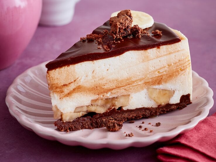 Ein Stück Schoko-Bananen-Torte mit Eierlikör auf einem rosa Teller auf lila-farbenem Hintergrund. 