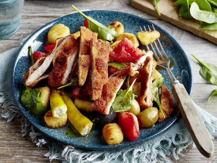 Antipasti-Schnitzel-Salat auf einem blauen Teller auf einem hellblauen Geschirrtuch auf hölzernem Hintergrund.