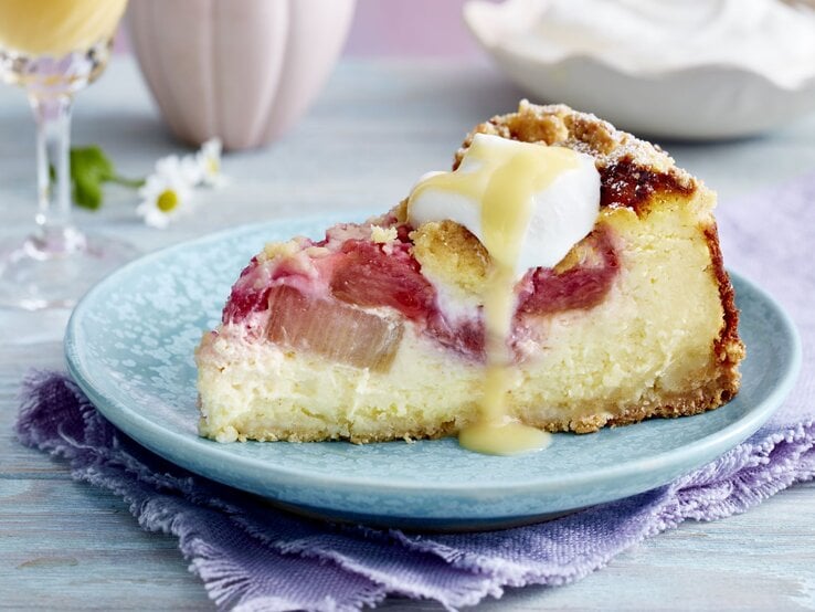 Rhabarber-Käsekuchen auf einem blauen Hintergrund. Hinten Gläser und Gänseblümchen verteilt.